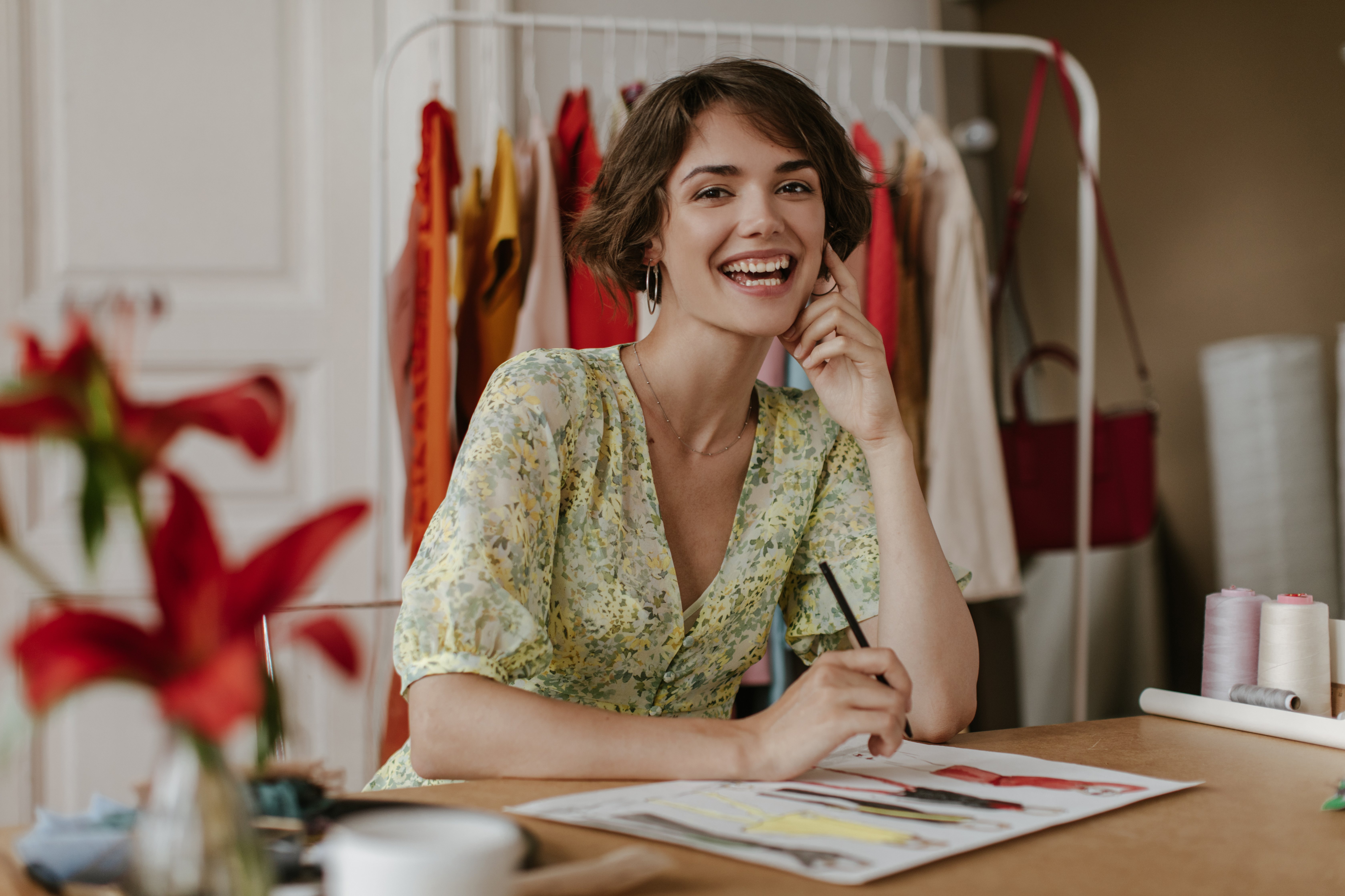 brown-eyed-curly-brunette-short-haired-woman-in-floral-trendy-dress-smiles-looks-at-camera-hoods-pencil-and-designs-new-clothes